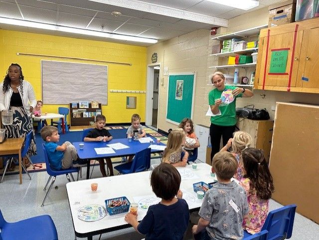 Teacher shows drawing to diverse group of children at table in brightly colored classroom.