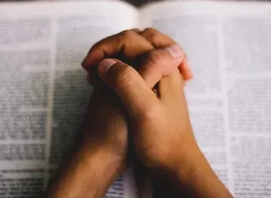 A person is praying with their hands folded over a bible.