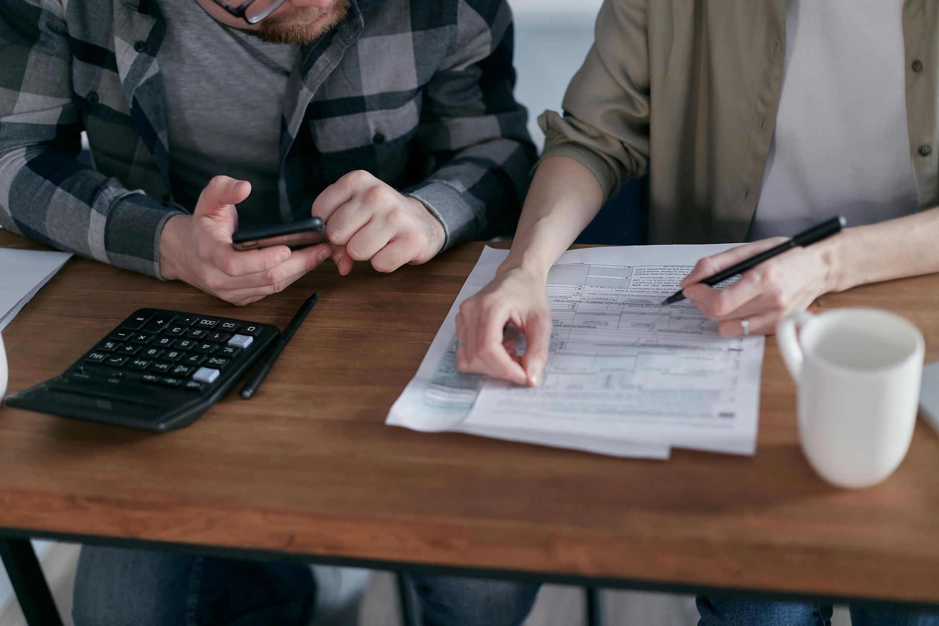 Dos personas revisando documentos con una calculadora y un teléfono inteligente sobre una mesa de madera.