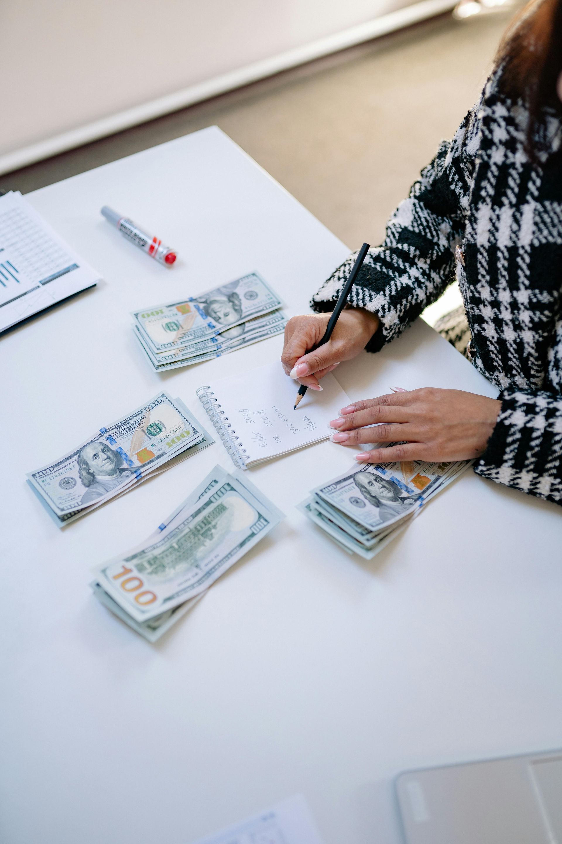 Mujer escribiendo a cuadros, fajos de billetes de 100 dólares sobre una mesa blanca.