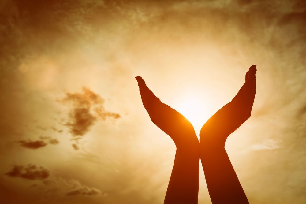 Hands cupped, silhouetted against a bright, golden sunset in a cloudy sky.