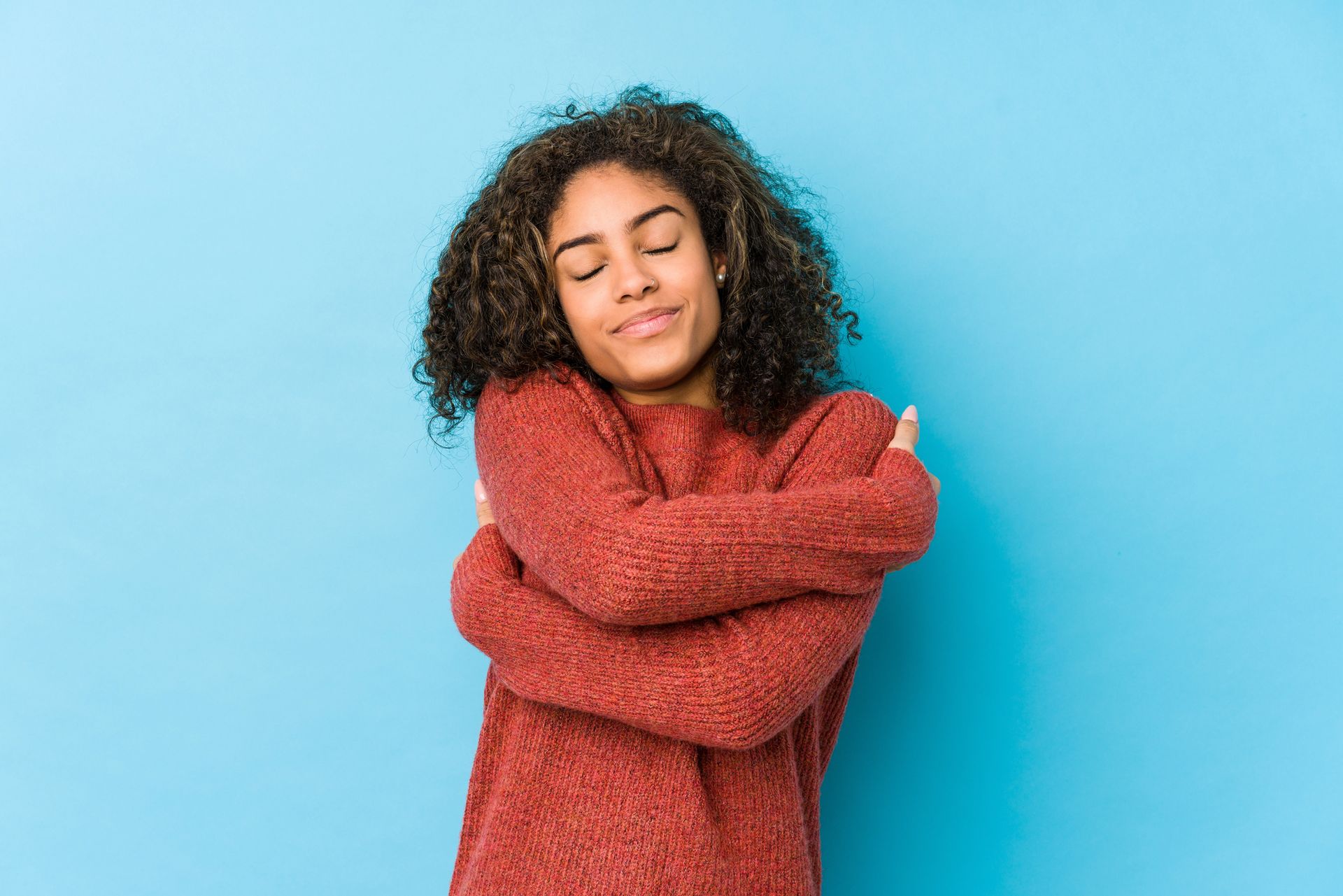 Young woman with curly hair embracing herself, smiling with eyes closed against a blue background.