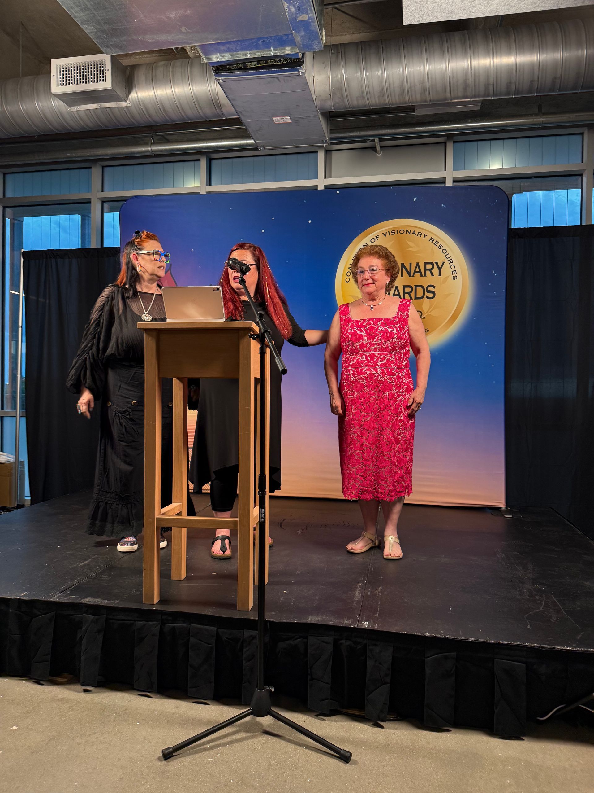 Three people on a stage near a podium, with a blue backdrop and circular event sign.