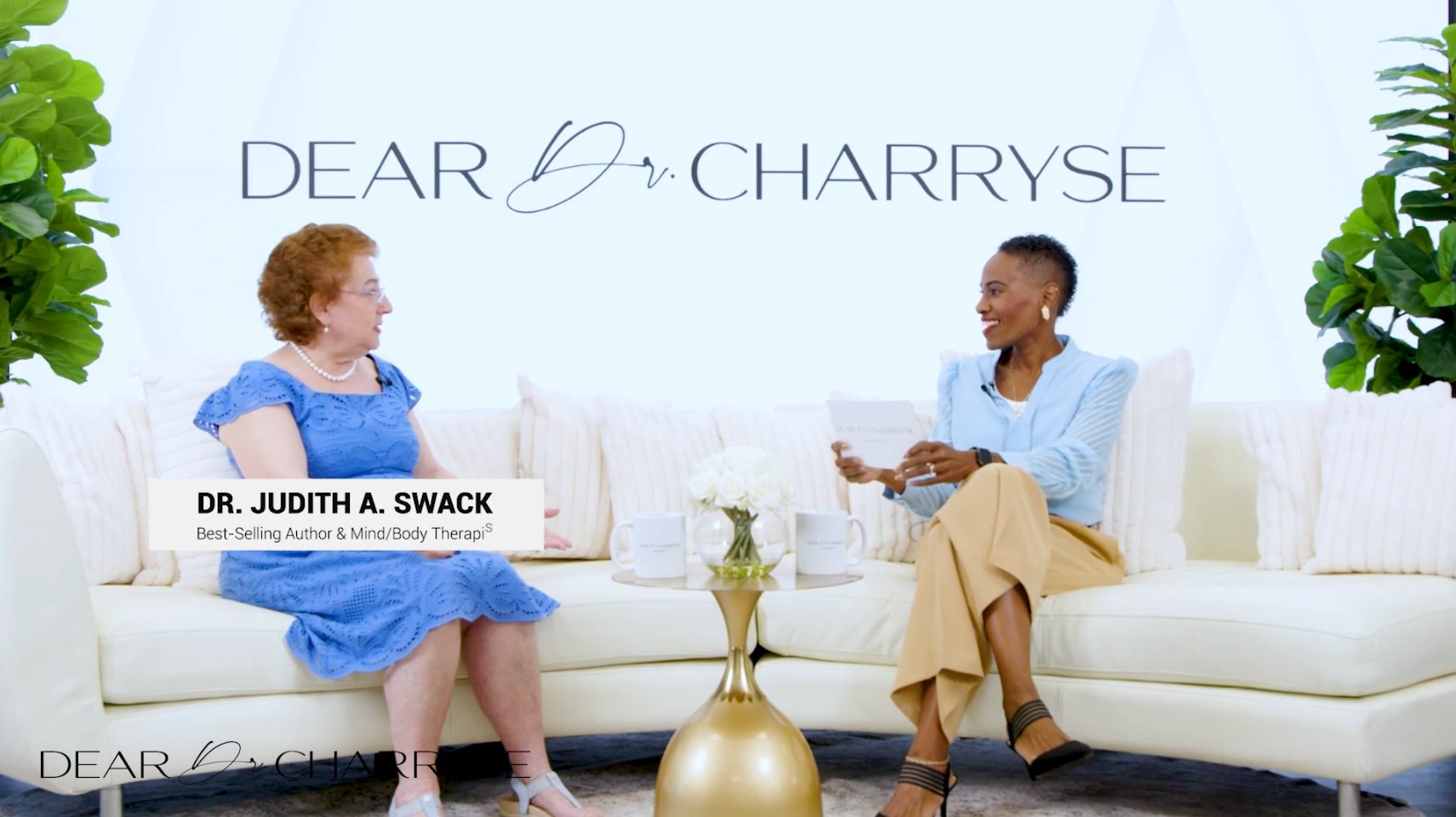 Woman in a blue dress and pearl necklace seated on a white couch, speaking in a bright living room.