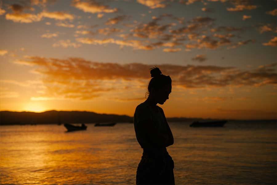 Silhouette of a person with arms crossed, gazing at a sunset over the water, boats in the distance.