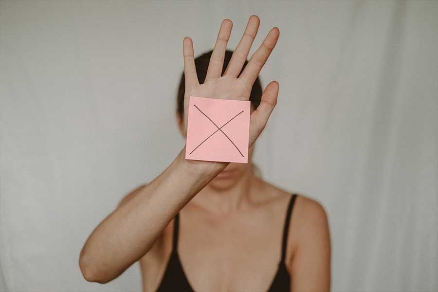 Person holding up hand, pink sticky note with black X covers face, blurred background.