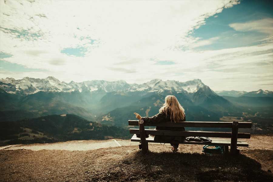 Woman with long blonde hair sits on a bench overlooking snow-capped mountains and a valley. Bright sky with clouds.