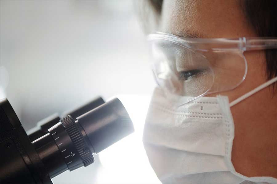 Person in lab goggles and mask looking through a microscope.