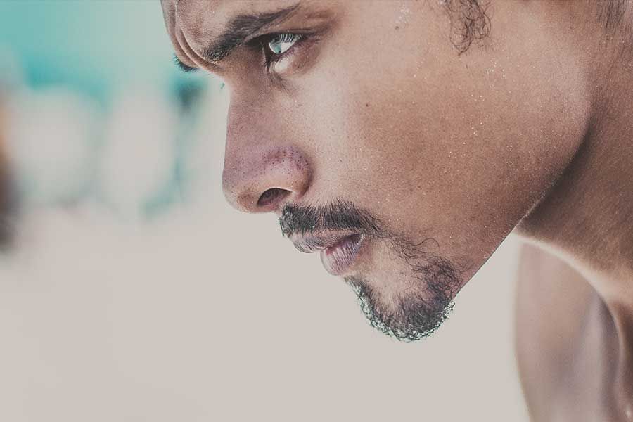 Man with focused expression, looking down. Sweating, short beard, outdoor setting, blurred background.
