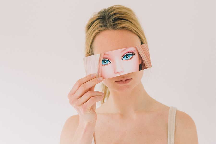 Woman holding a Barbie doll face cutout over her eyes against a white background.