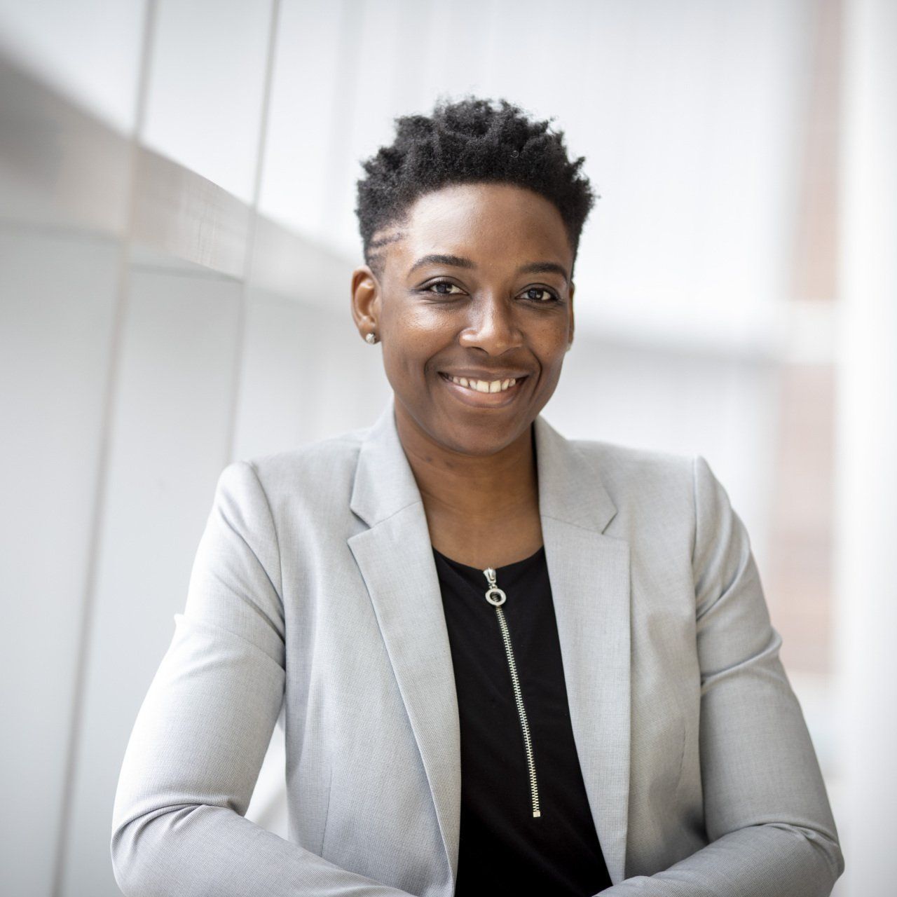Woman with short hair, smiling in a light gray blazer and black shirt, by a window.