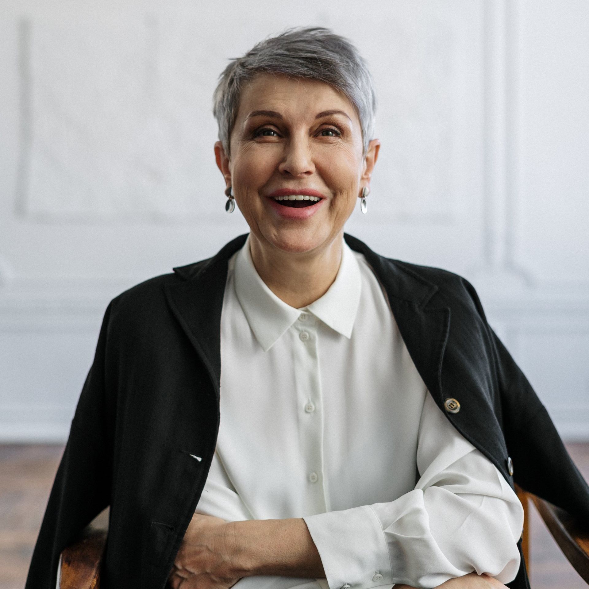 Woman with short gray hair, smiling, wearing a white blouse and black blazer in a bright room.