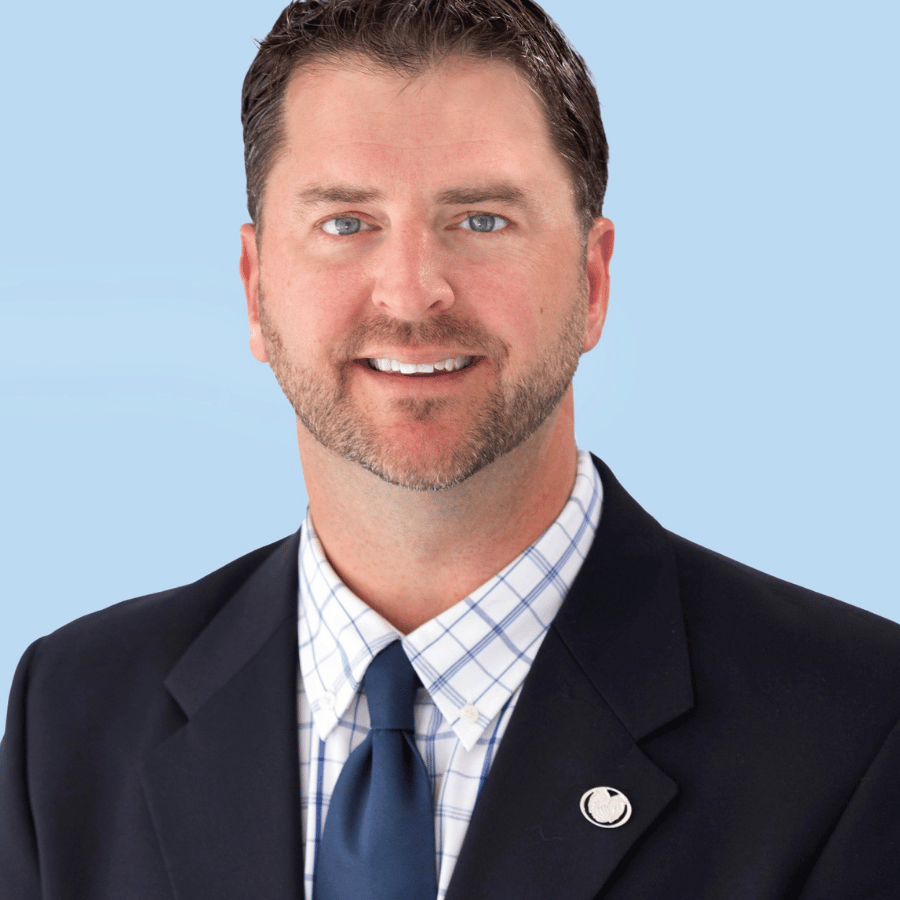 Professional headshot of a smiling man in a dark suit and blue tie on a light blue background