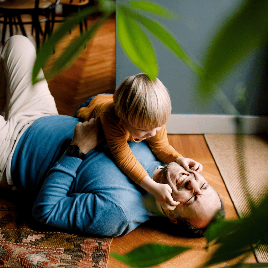 Child playing on top of an adult on a hardwood floor, with green leaves in the foreground