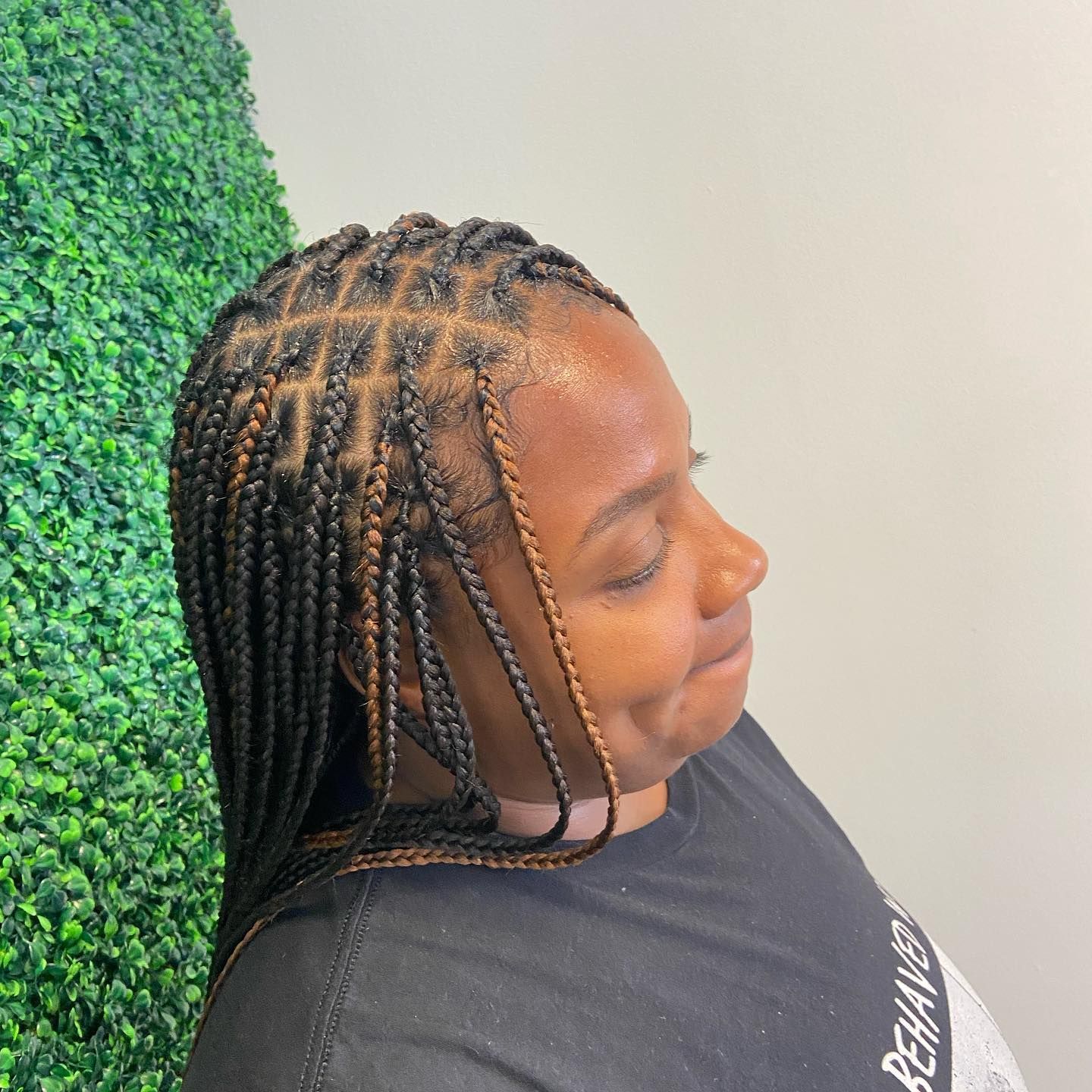 Person with box braids next to a green wall; wearing a black shirt with text.