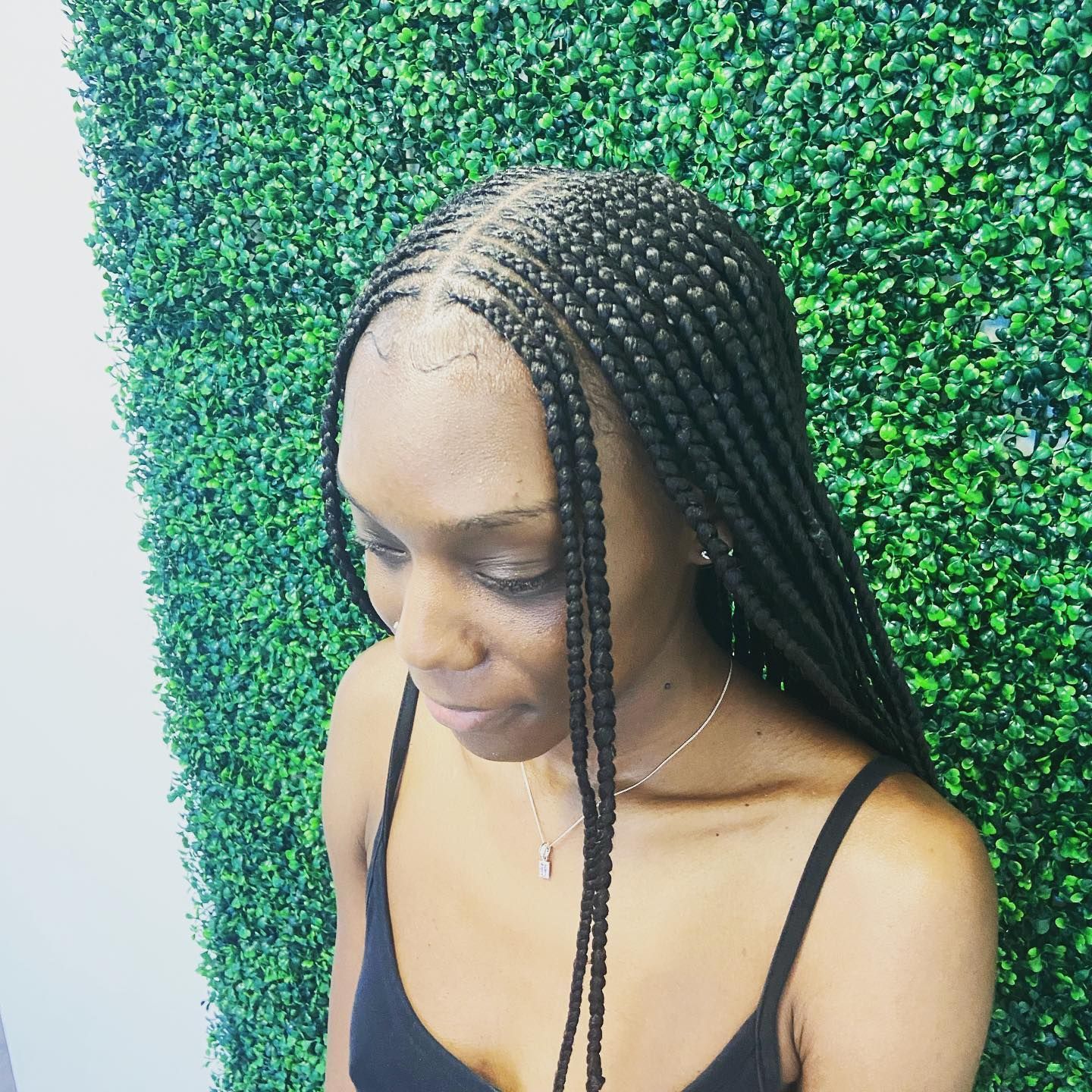 Woman with long braided hair, wearing a black tank top, in front of a green wall.