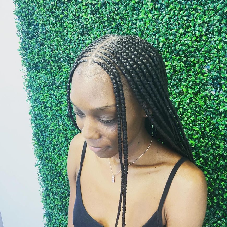 Woman with long braided hair, wearing a black tank top, in front of a green wall.