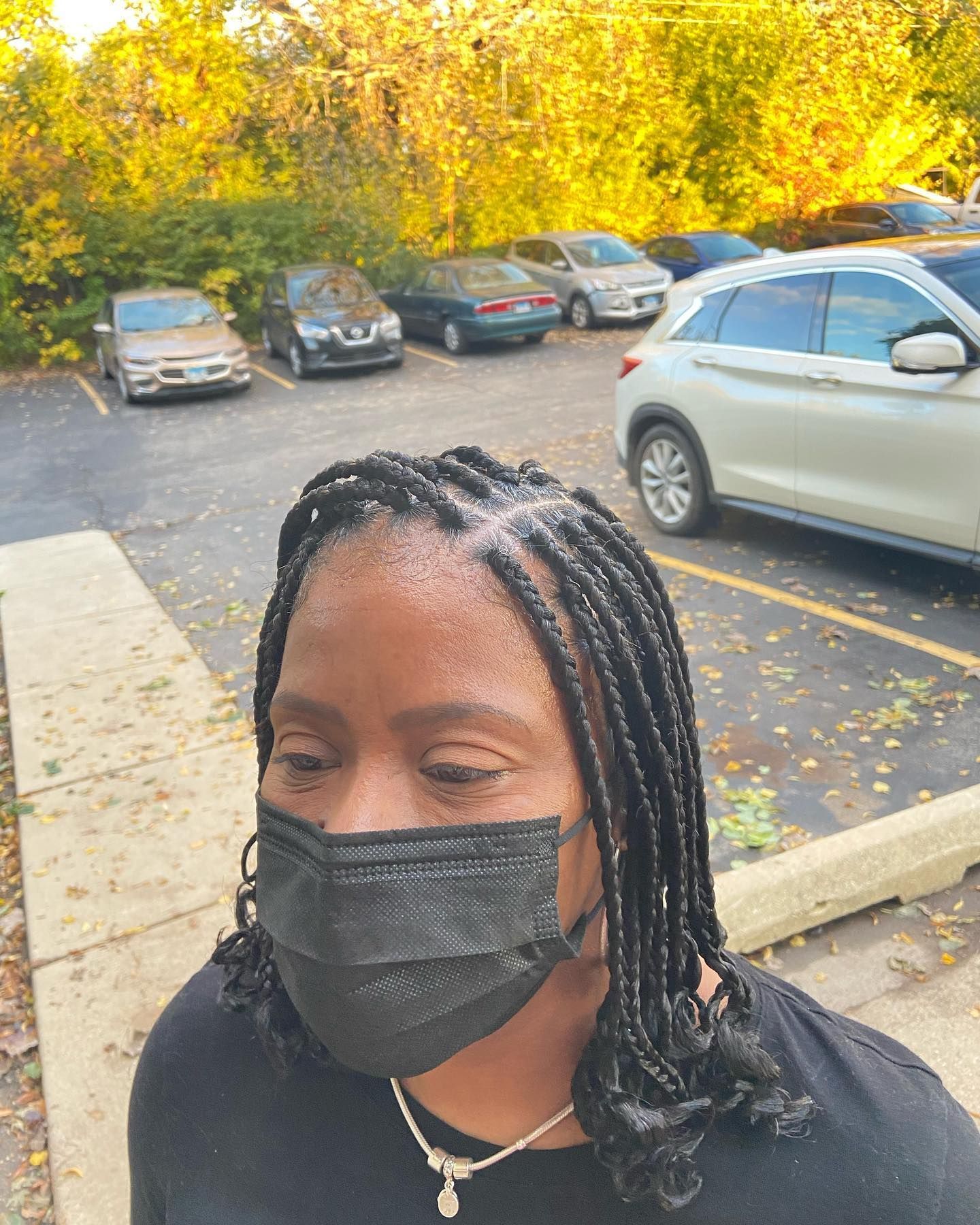 Woman with braided hair wearing a black face mask, in a parking lot. Cars and trees in the background.