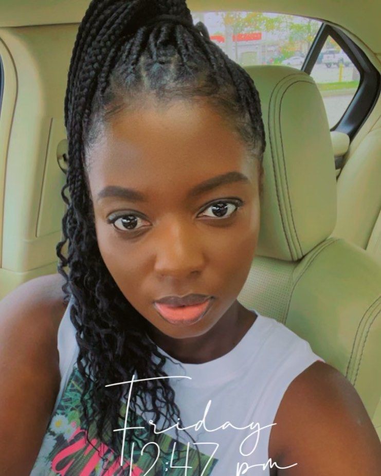 Woman in a car with braided hair, looking at the camera. She's wearing a white top.