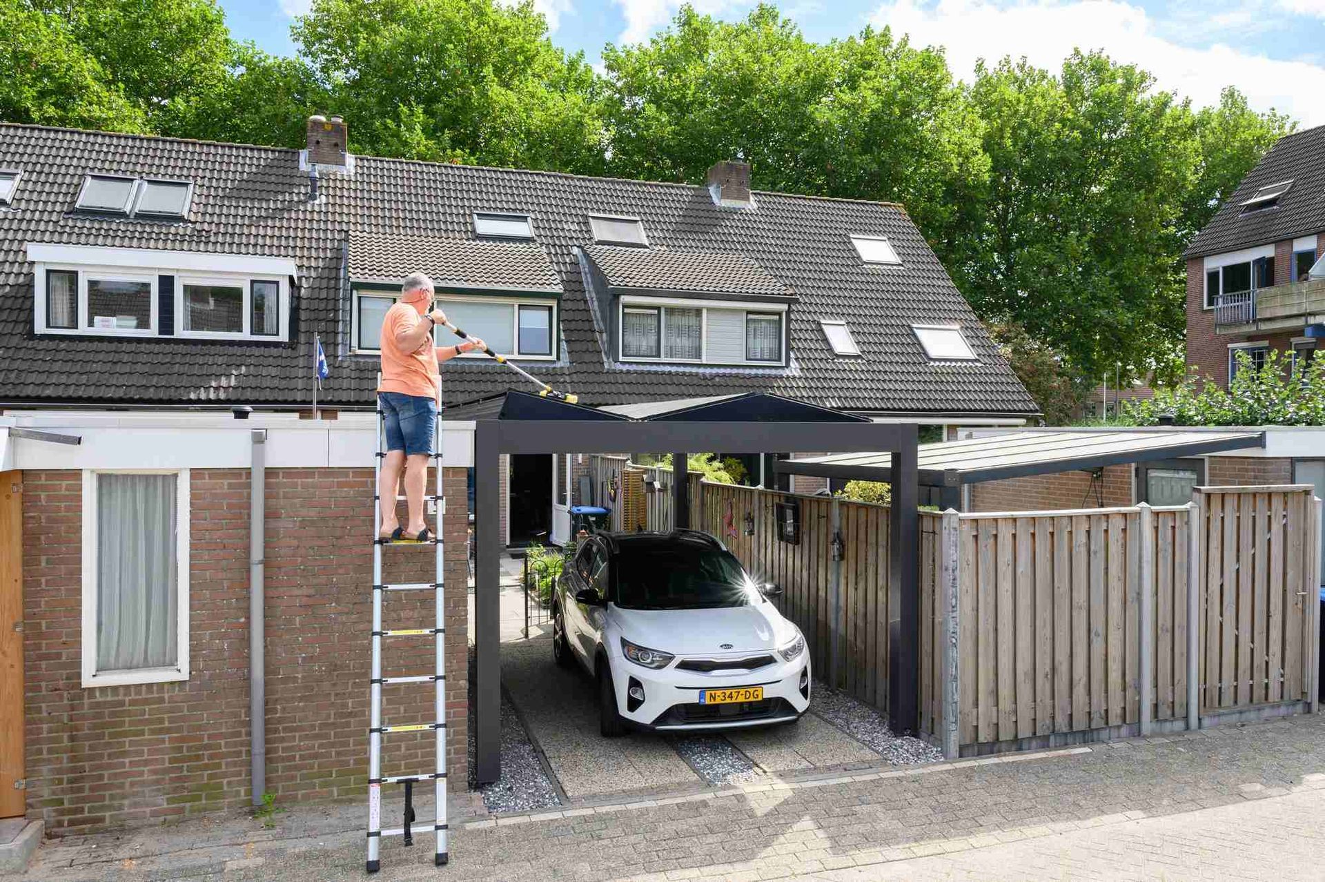 Een man staat op een ladder naast een auto onder een carport.