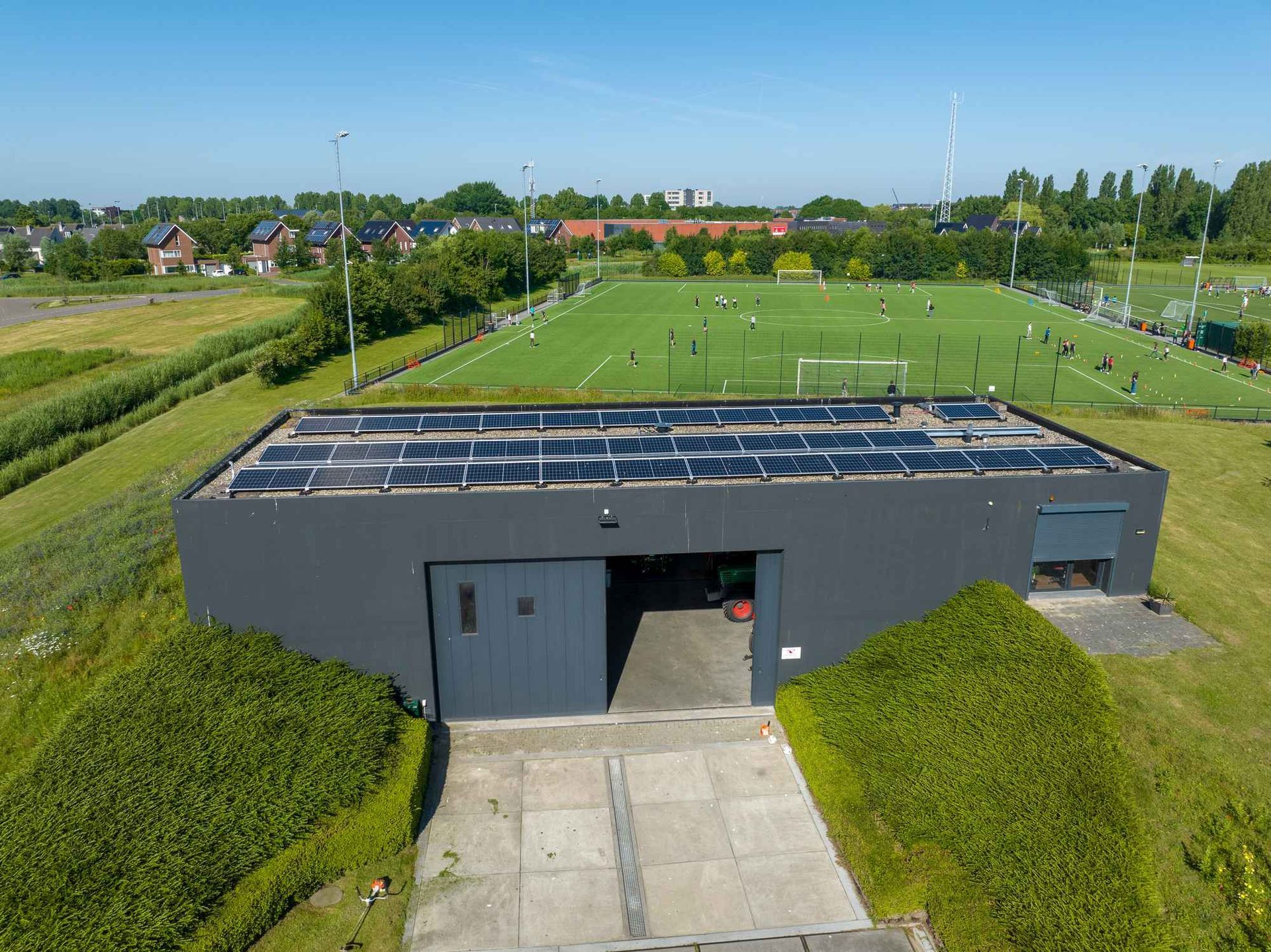 Een luchtfoto van een gebouw met zonnepanelen op het dak en een voetbalveld op de achtergrond.