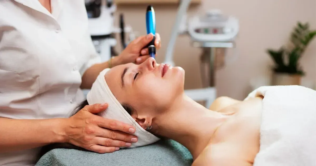 Spa esthetician applying treatment to a woman's face with a brush while she lies on a treatment bed.