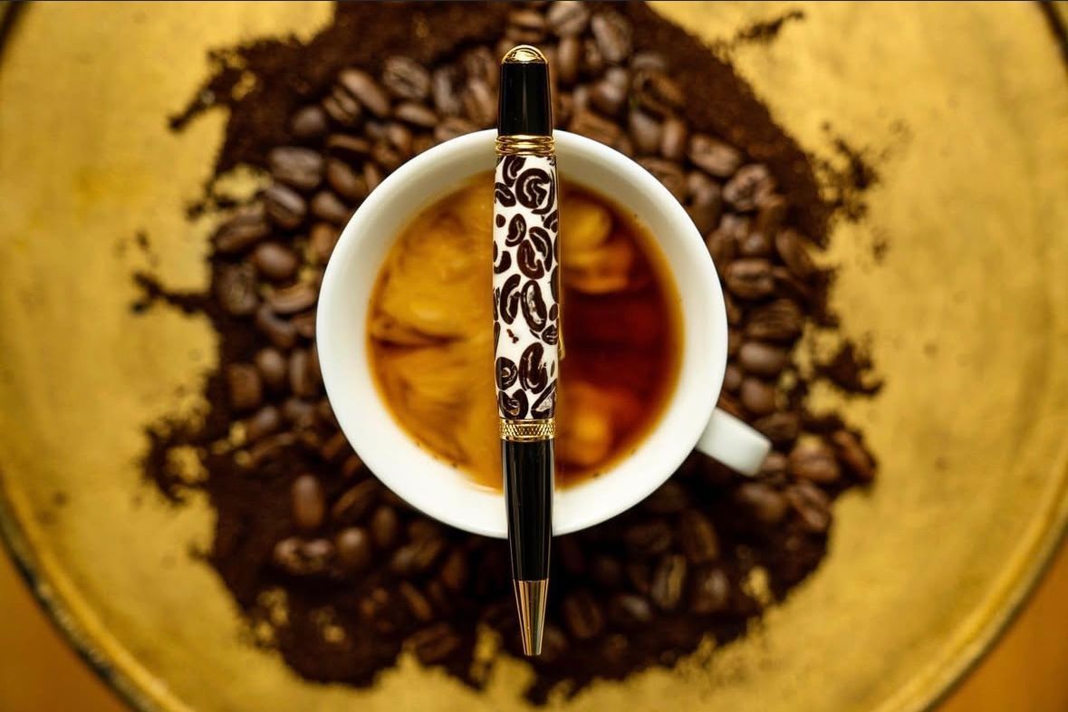 A patterned pen rests vertically across a cup of coffee, surrounded by coffee beans on a golden plate.