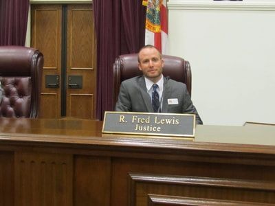 A man in a suit and tie is sitting at a desk with a name plate that says r. fred lewis justice