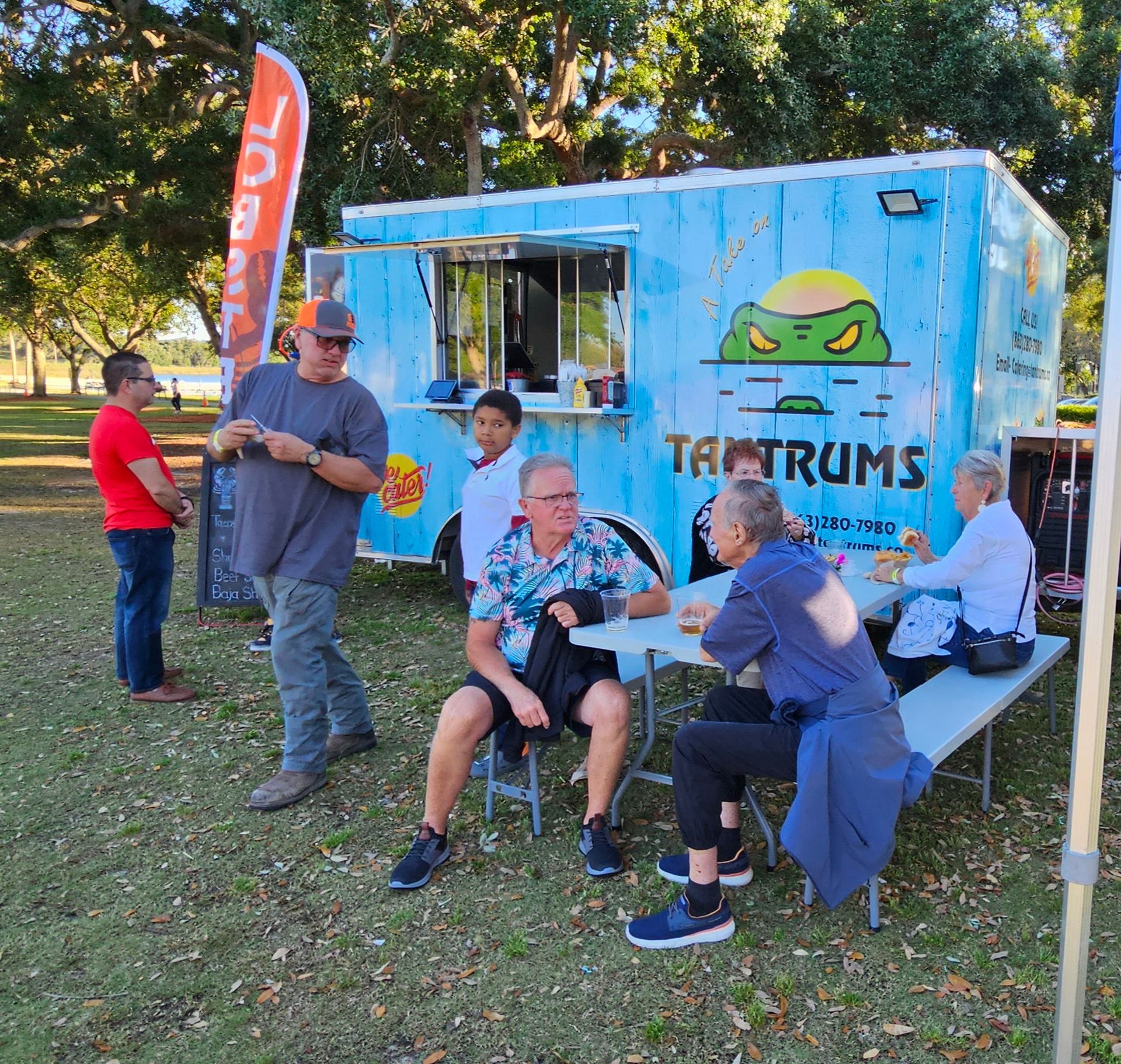 A group of people sit at tables in front of a food truck that says tamtrums