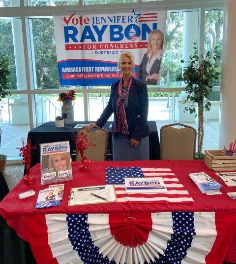 A woman stands in front of a table with a sign that says vote jennifer raybon