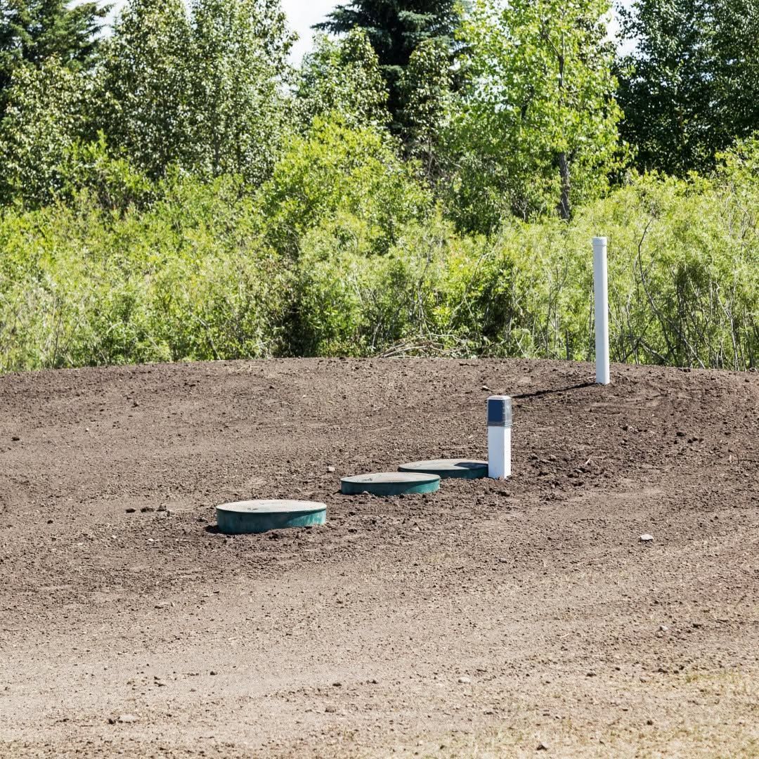 Outdoor dirt area with three green utility covers and a white pipe marker near trees