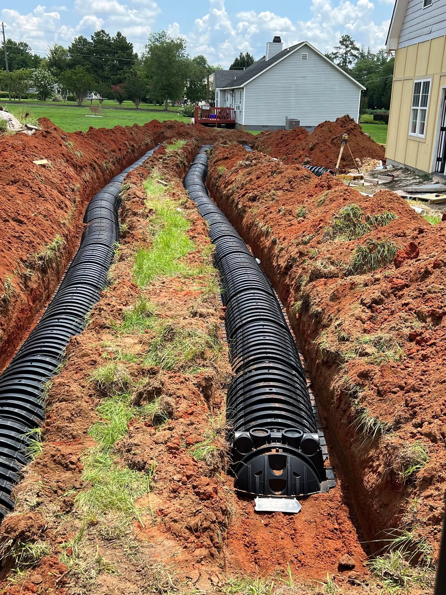 Trench with black drainage pipes laid in red soil beside a house under construction