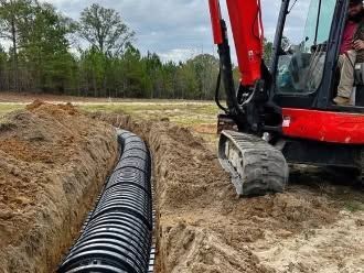 Red excavator beside a trench with a black corrugated pipe being laid in a muddy field