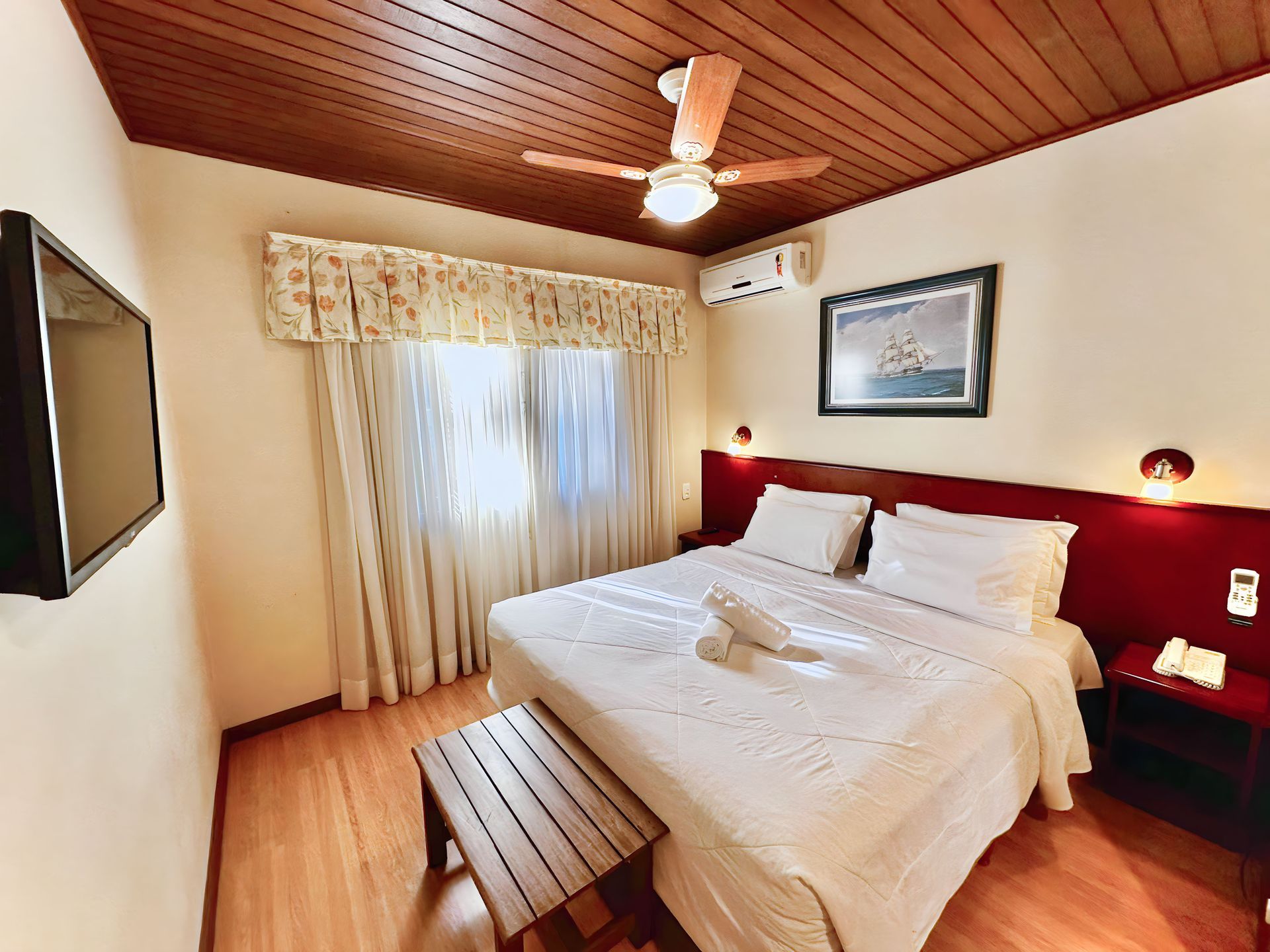 Um quarto de hotel com cama, ventilador de teto, televisão e banco.