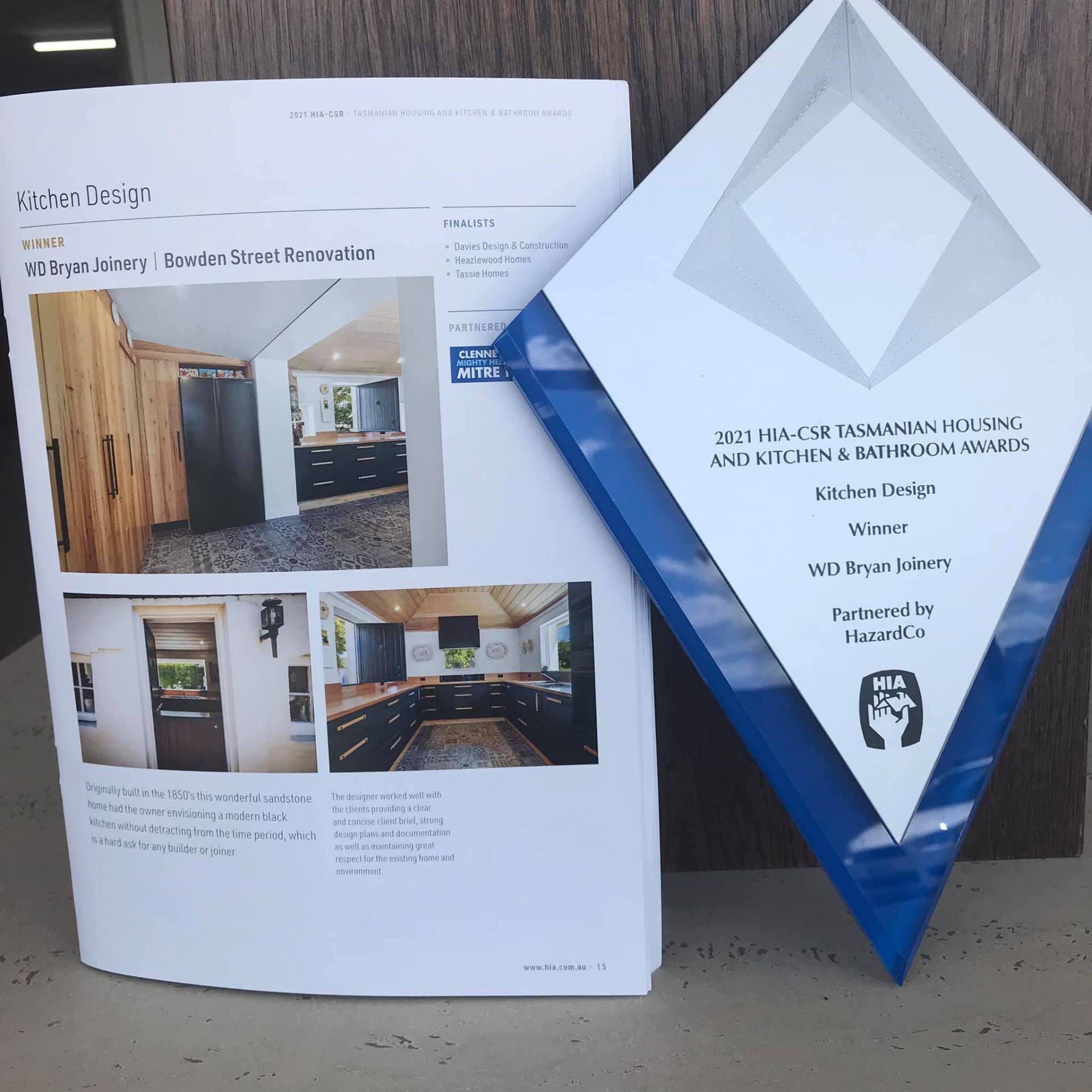 Kitchen Design Winner 2021 — Glenorchy, TAS — WD Bryan Joinery House Brochure and blue award plaque on a wooden table, showing interior renovation photos and text