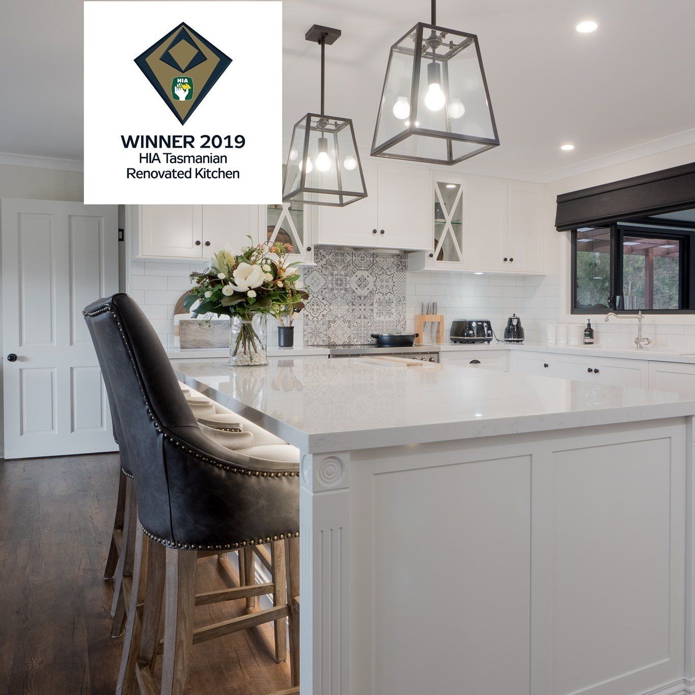 Bright white kitchen with island, black stools, pendant lights, and hardwood floors; award badge in corner