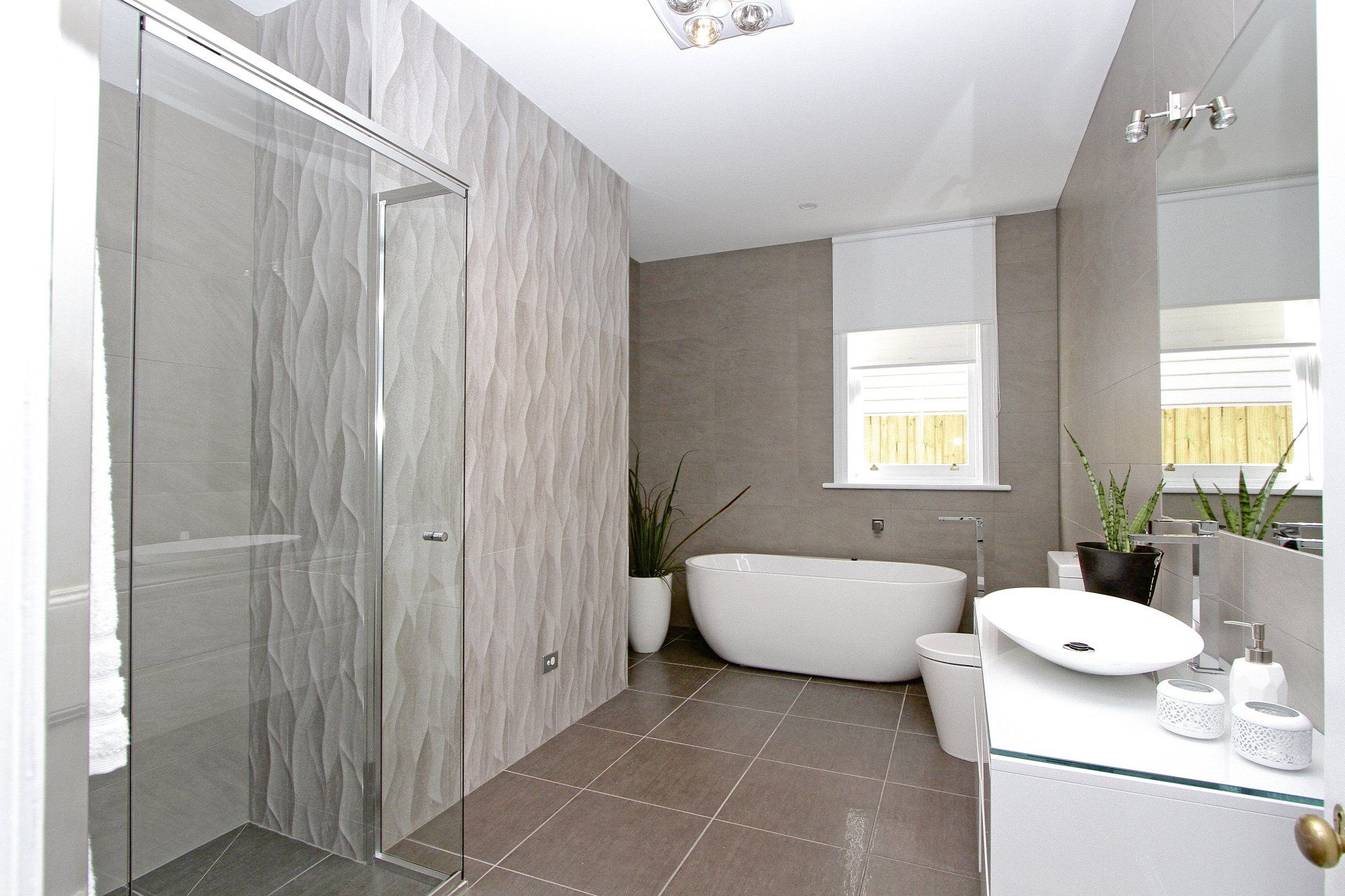 Modern gray bathroom with glass shower, freestanding tub, and vanity sink near windows
