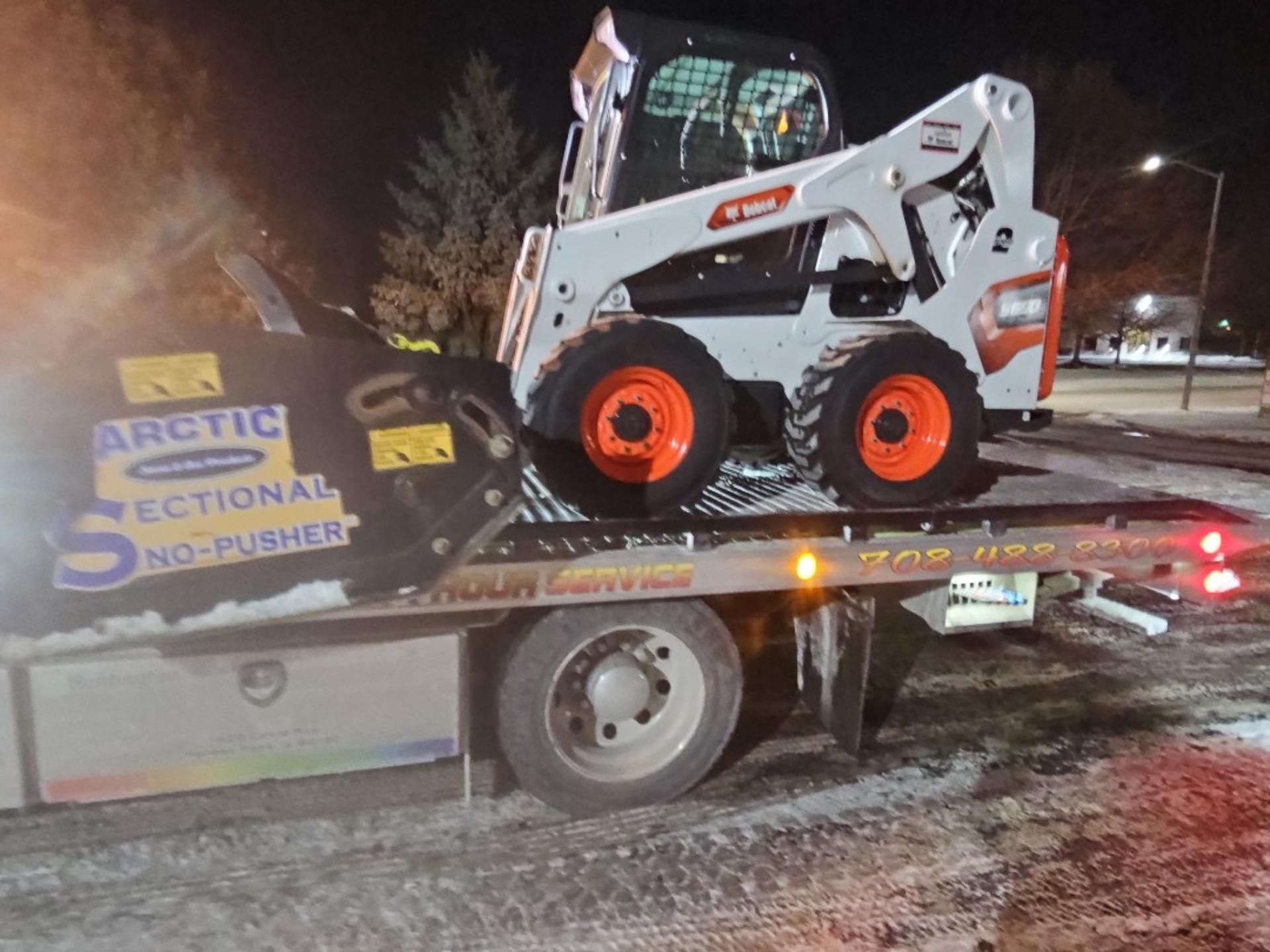White skid-steer loader on a flatbed trailer at night, with an Arctic Sectional snow pusher nearby.