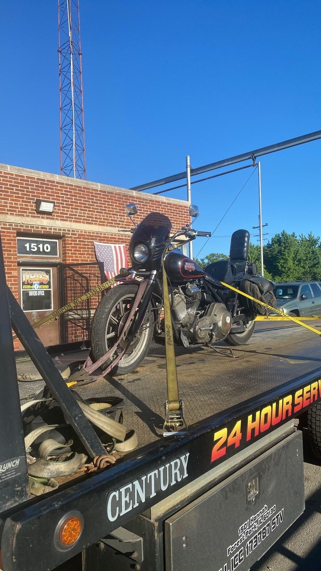 Motorcycle secured on a flatbed tow truck outside a brick building on a sunny day.