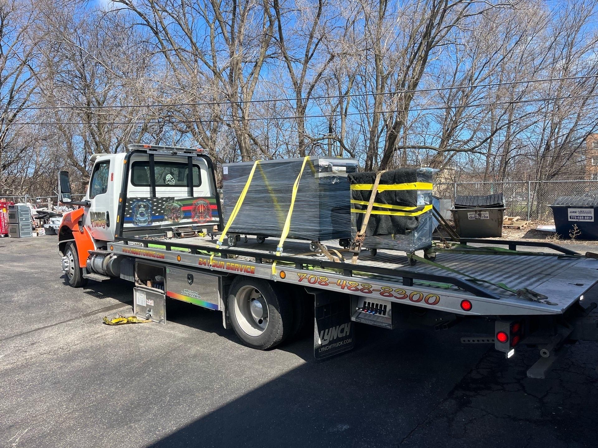 Tow truck carrying strapped equipment on a flatbed in a parking lot with bare trees nearby