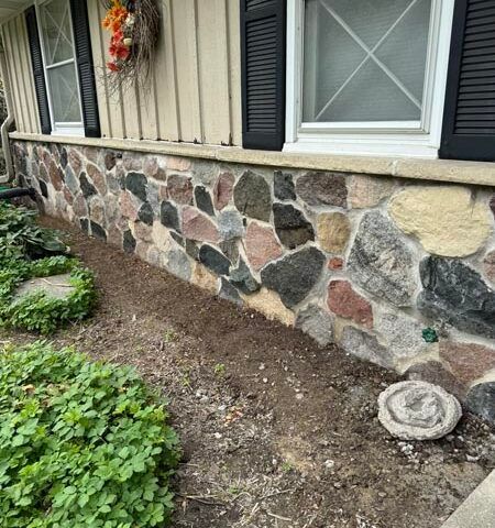 Stone foundation and beige siding of a house with black shutters, flower wreath, and a mulch bed in front.