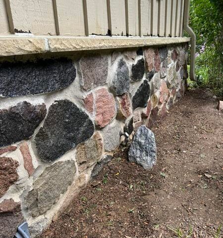 Stone foundation wall beside a dirt path, with beige siding above and trees in the background.