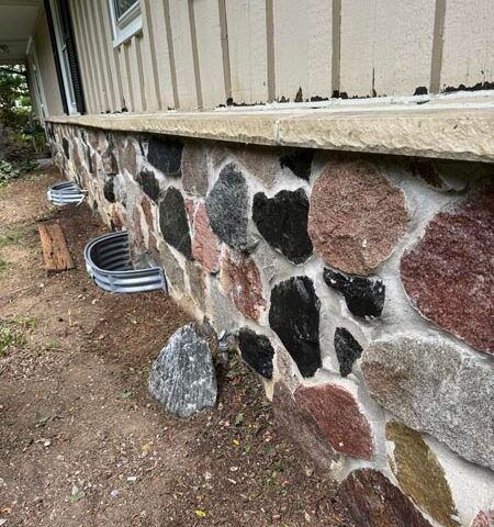 Stone-faced foundation wall beside a house with a dirt yard and coiled drainage pipe at the base