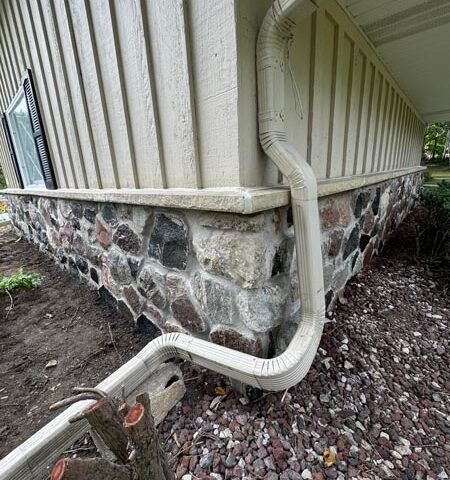 House corner with beige siding, stone foundation, and a downspout draining into a gutter extension over gravel.