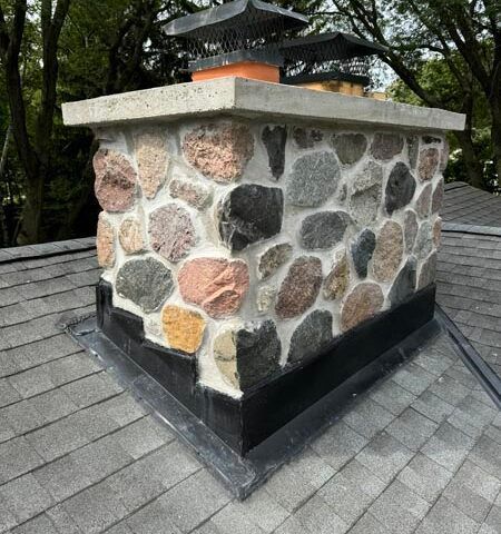Stone chimney with black trim on a shingled roof, topped by a metal cap, surrounded by trees.
