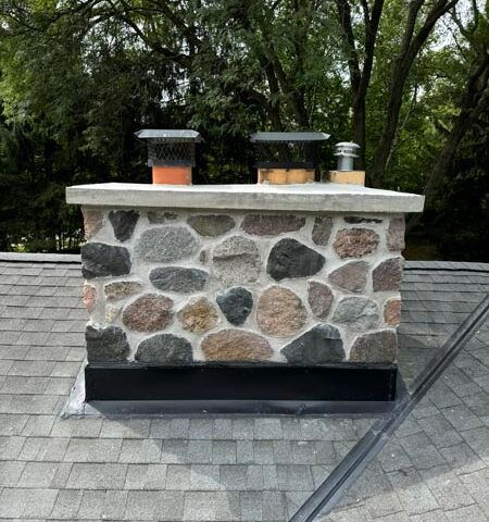 Stone chimney with black cap on a gray shingled roof, surrounded by trees.