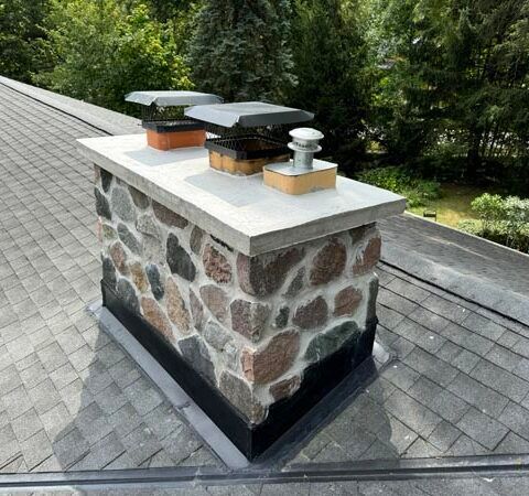 Stone chimney on a shingled roof with two metal vent caps, surrounded by green trees.