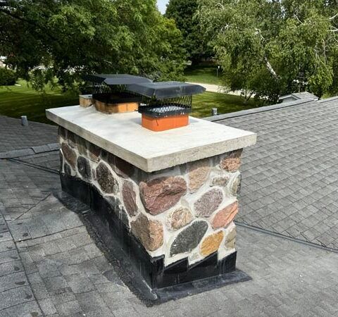 Stone outdoor chimney on a gray shingled roof with black caps, surrounded by green trees.