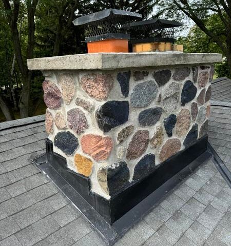 Stone chimney on a gray shingle roof with a black cap and forested background