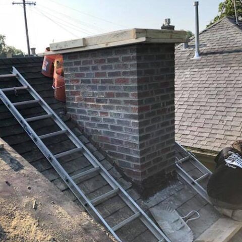 Brick chimney on a roof with two ladders leaning against it and two red fire extinguishers mounted on the side.